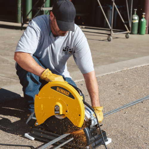 A man in a gray shirt and black cap uses a yellow DeWalt metal chop saw, cutting metal, with sparks flying. He stands on a paved surface outdoors.