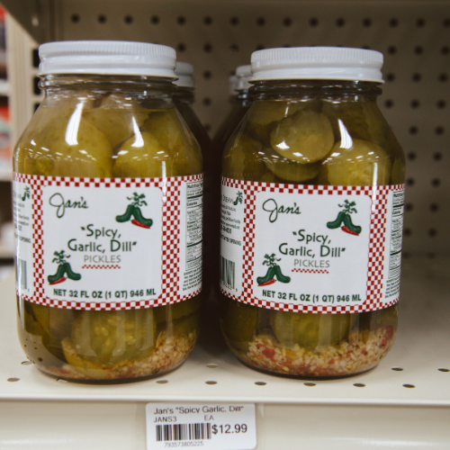 Two jars of Jan's "Spicy Garlic Dill" pickles on a store shelf, featuring green pickles and spices visible through clear glass. Price tag reads $12.99.
