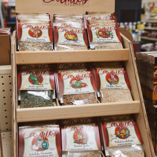 A store display features various packets of Carrie's Kitchen dip and dressing mixes on wooden shelves, showcasing colorful packaging with vegetable images.