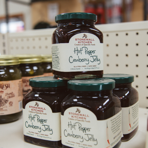 Four jars of "Hot Pepper Cranberry Jelly" with green lids are stacked on a white retail shelf. The background shows blurred store shelves.