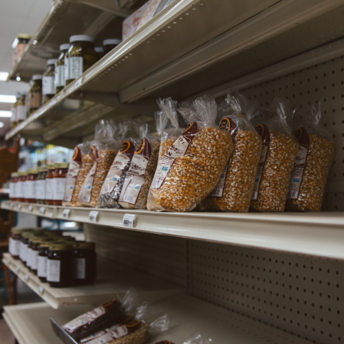Bags of popcorn kernels neatly arranged on a grocery store shelf, next to jars of preserves. The aisle is well-lit, creating a calm shopping atmosphere.