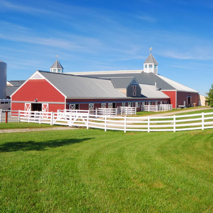 Farm SuppliesA large red barn with white trim and twin cupolas sits on a lush green lawn under a clear blue sky. A white fence surrounds the peaceful rural scene.